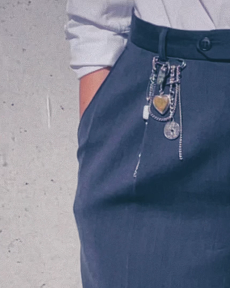 Person wearing a white shirt and grey pleated pants adorned by a safety pin with multiple charms, coins and objects attached to it.